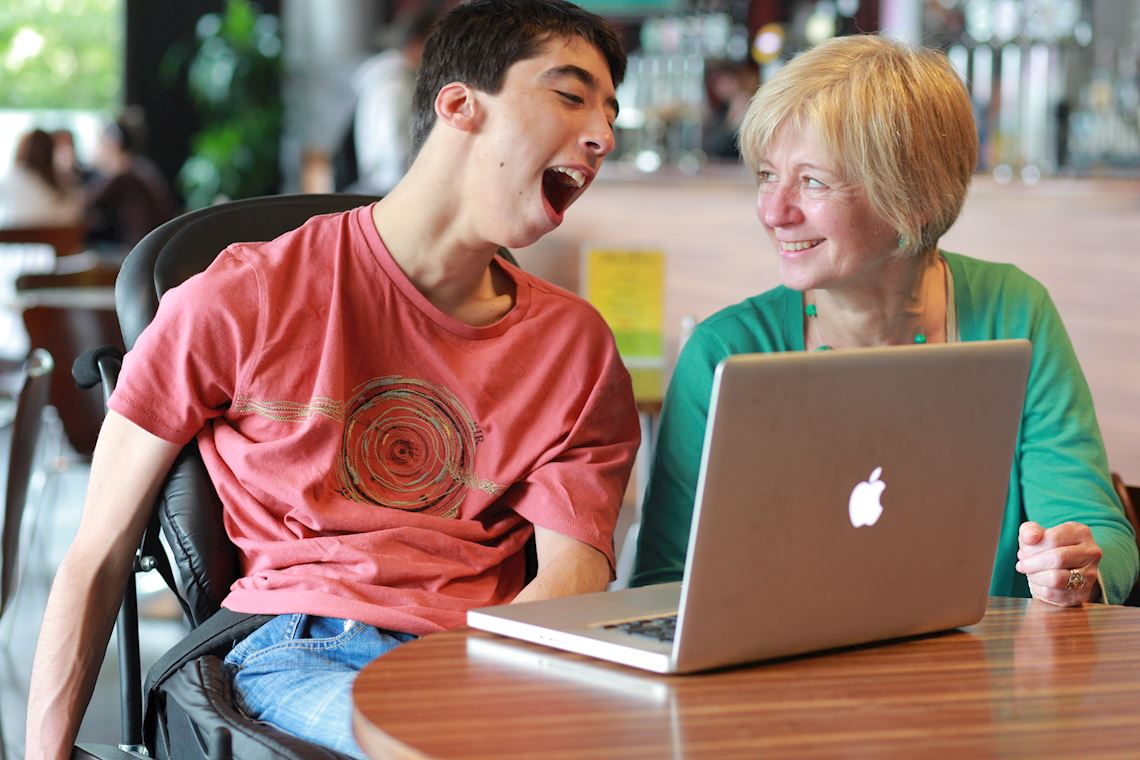 A woman helps a boy in a wheelchair look at a laptop