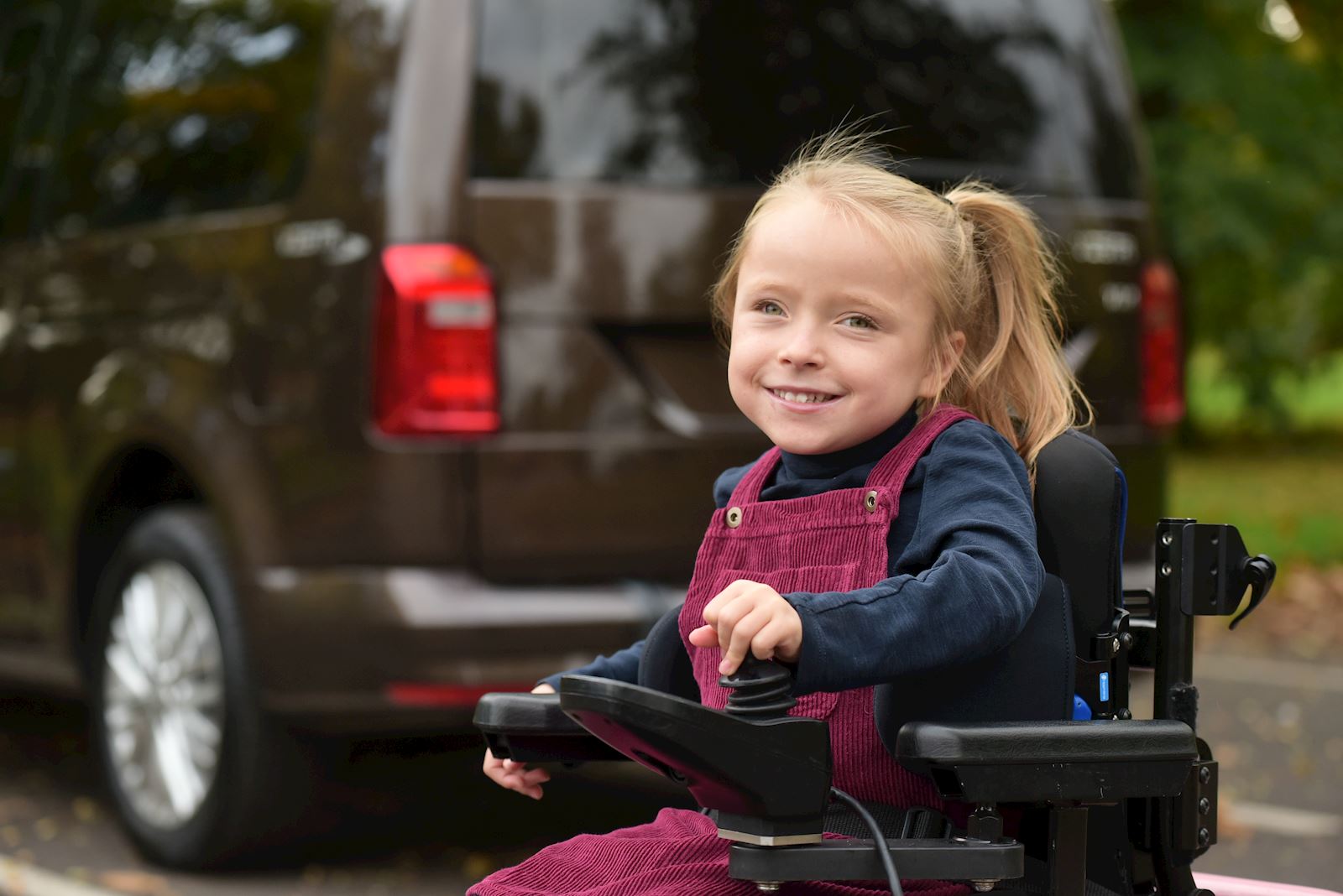A little girl in a powered wheelchair, smiling, with a WAV in the background