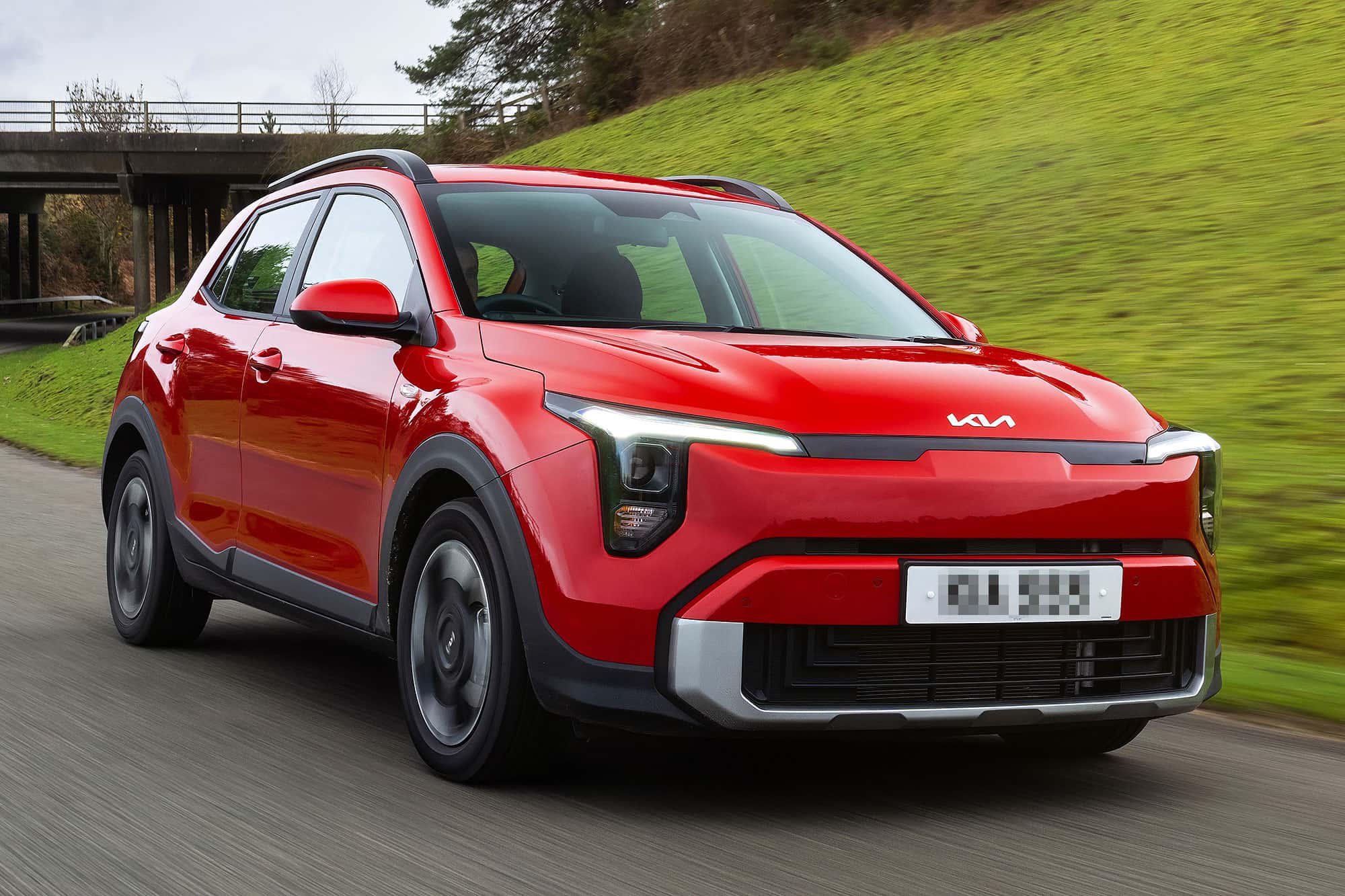 Red Kia crossover driving on a country road with green hillside and bridge in the background.