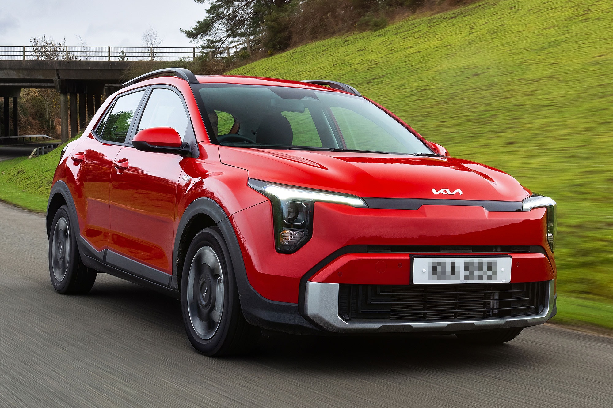 Red Kia crossover driving on a country road with green hillside and bridge in the background.