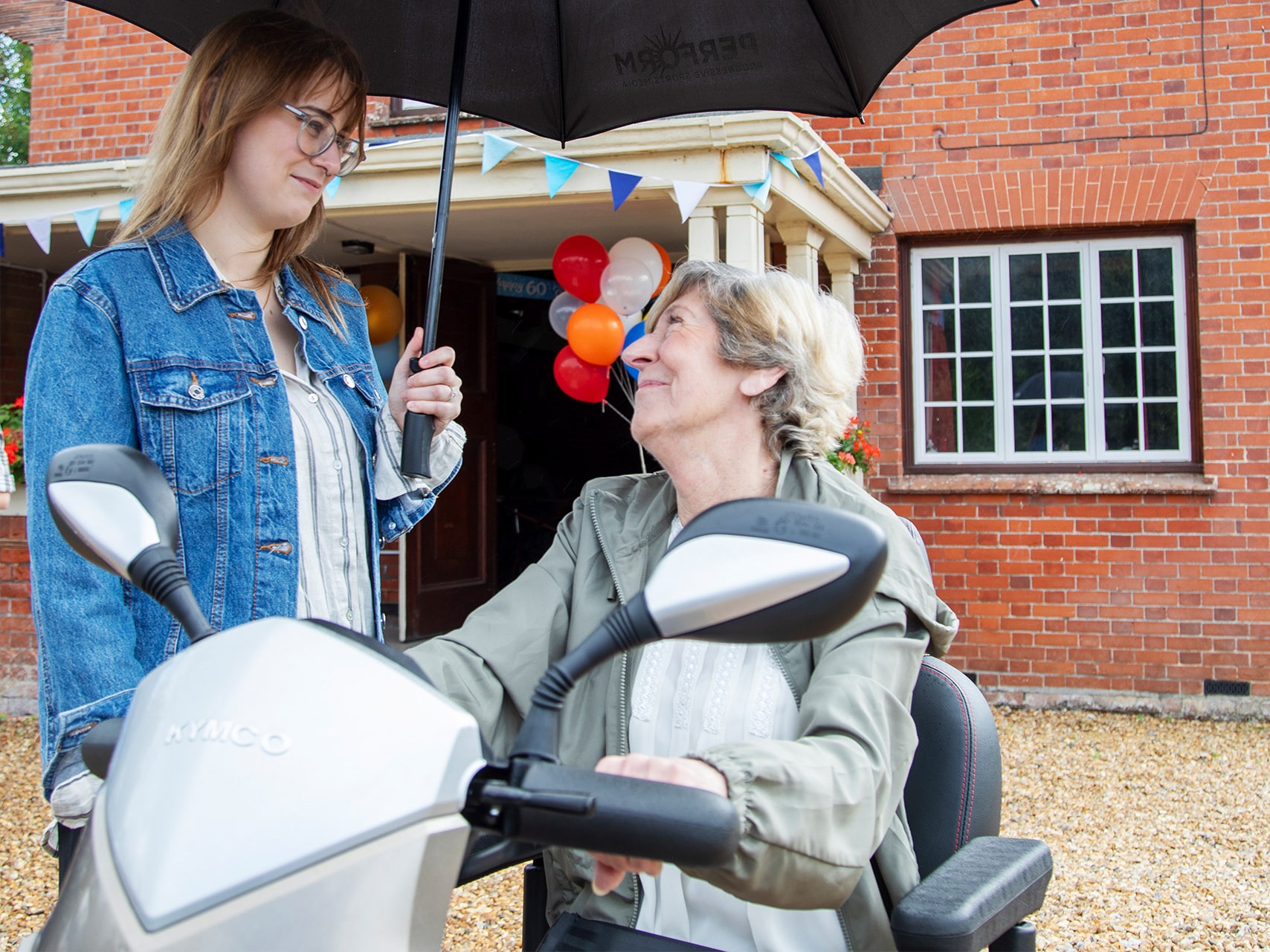 Young woman in denim jacket holds umbrella over older woman in mobility scooter outside red brick house with bunting.