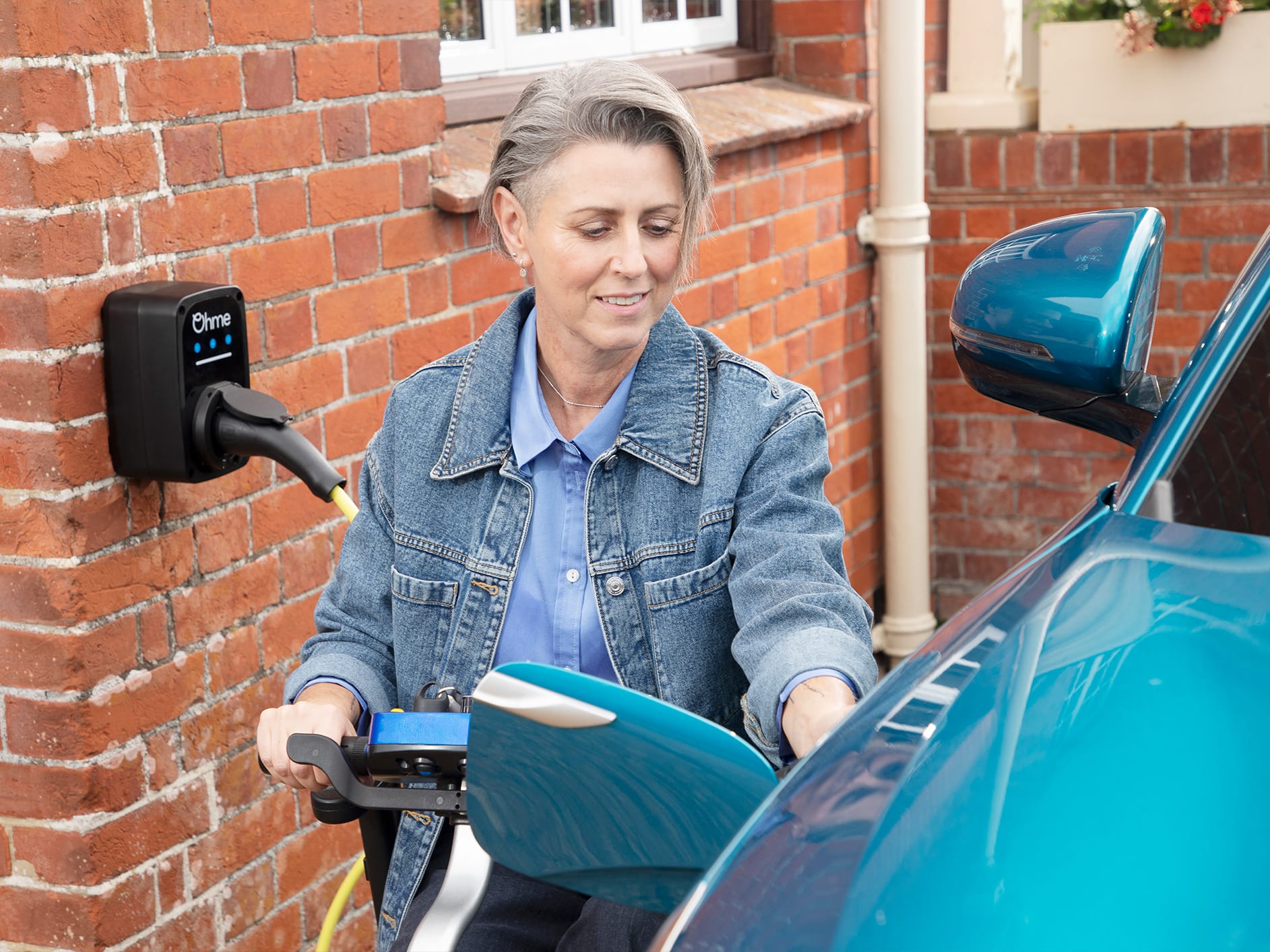 Woman in denim jacket charging blue electric car at home wall-mounted charging point on red brick wall