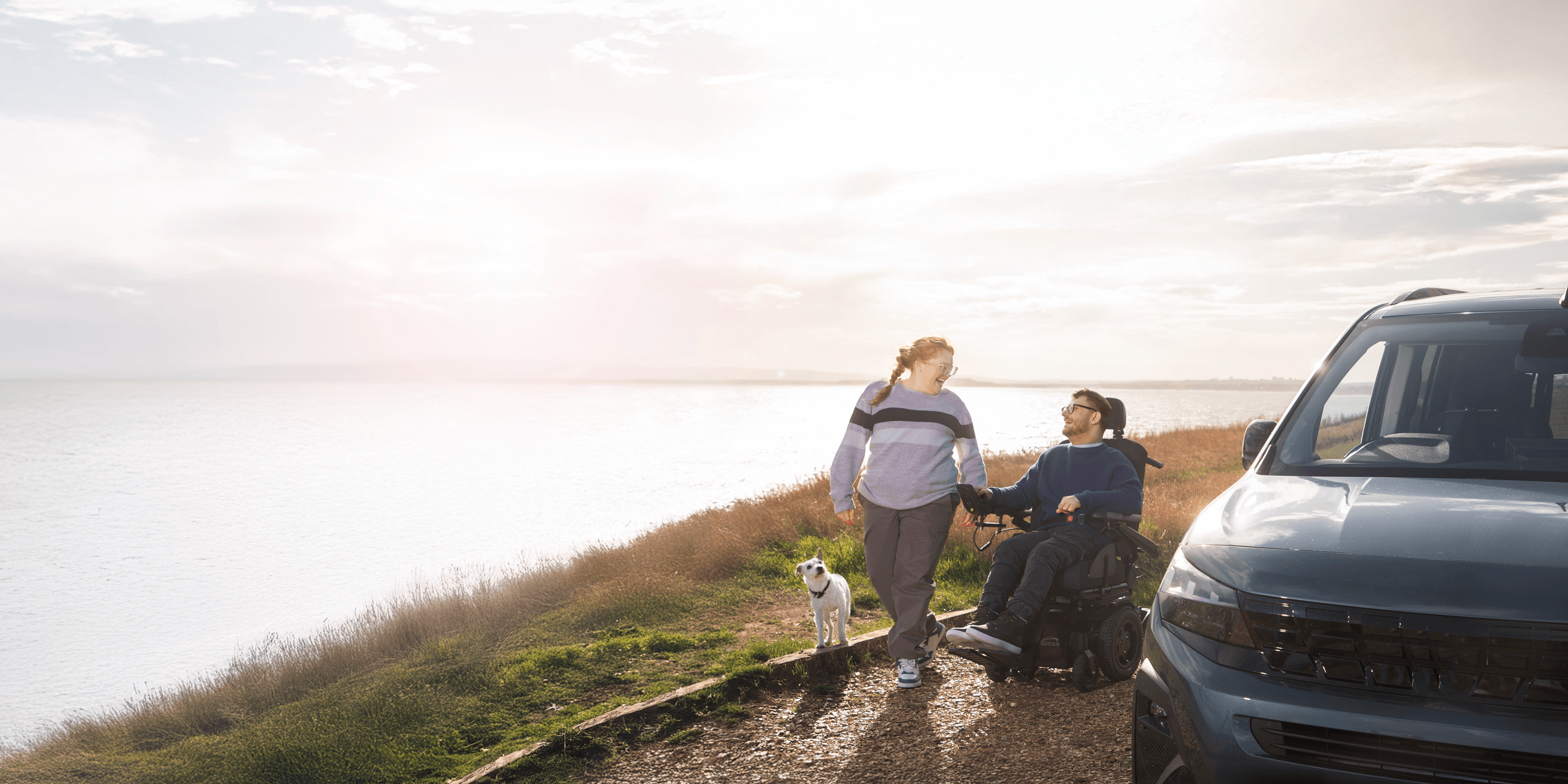 A man in a wheelchair and a woman walk a dog along a coastal path beside a blue Wheelchair Accessible Vehicle.