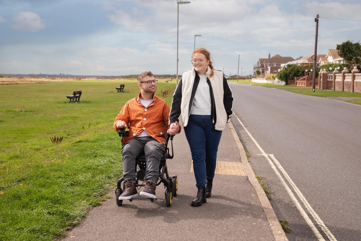 Young woman walking alongside man in wheelchair on coastal path, holding hands, with green grass and houses visible.