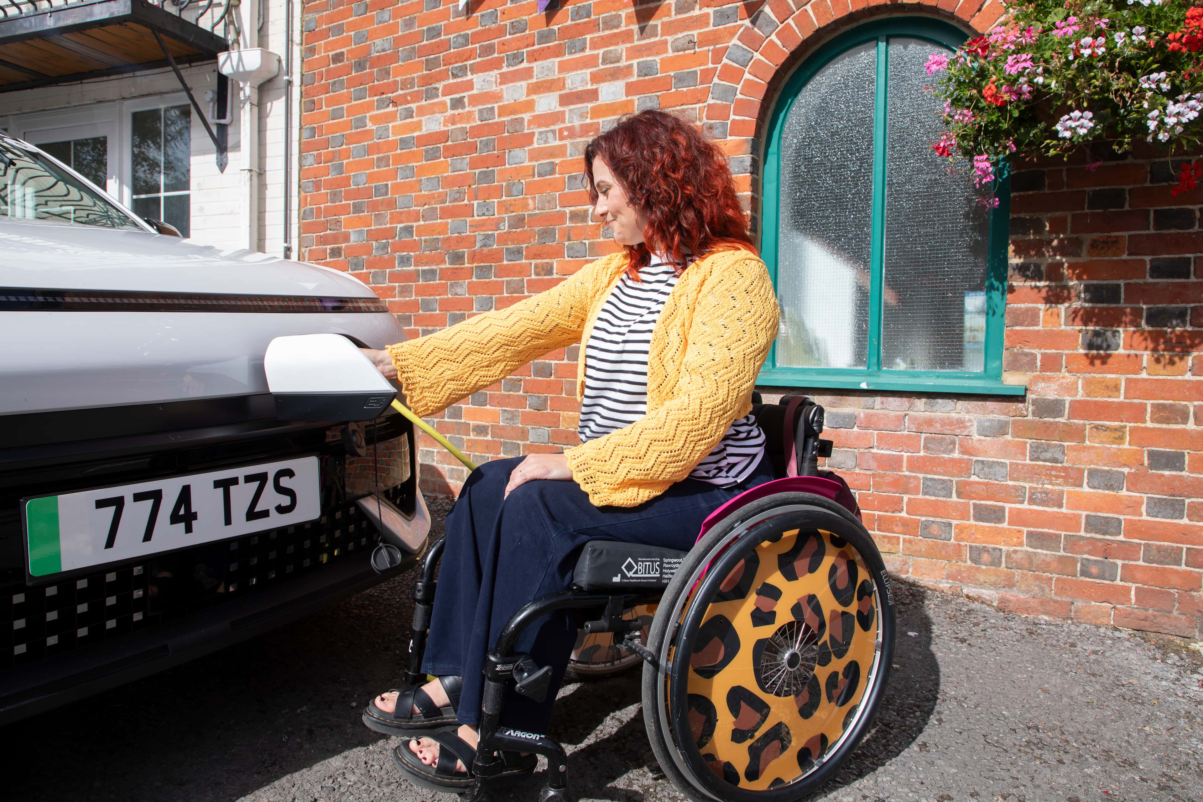 A woman in a manual wheelchair wearing a yellow cardigan and striped top charges a car outside a brick house with hanging flowers.