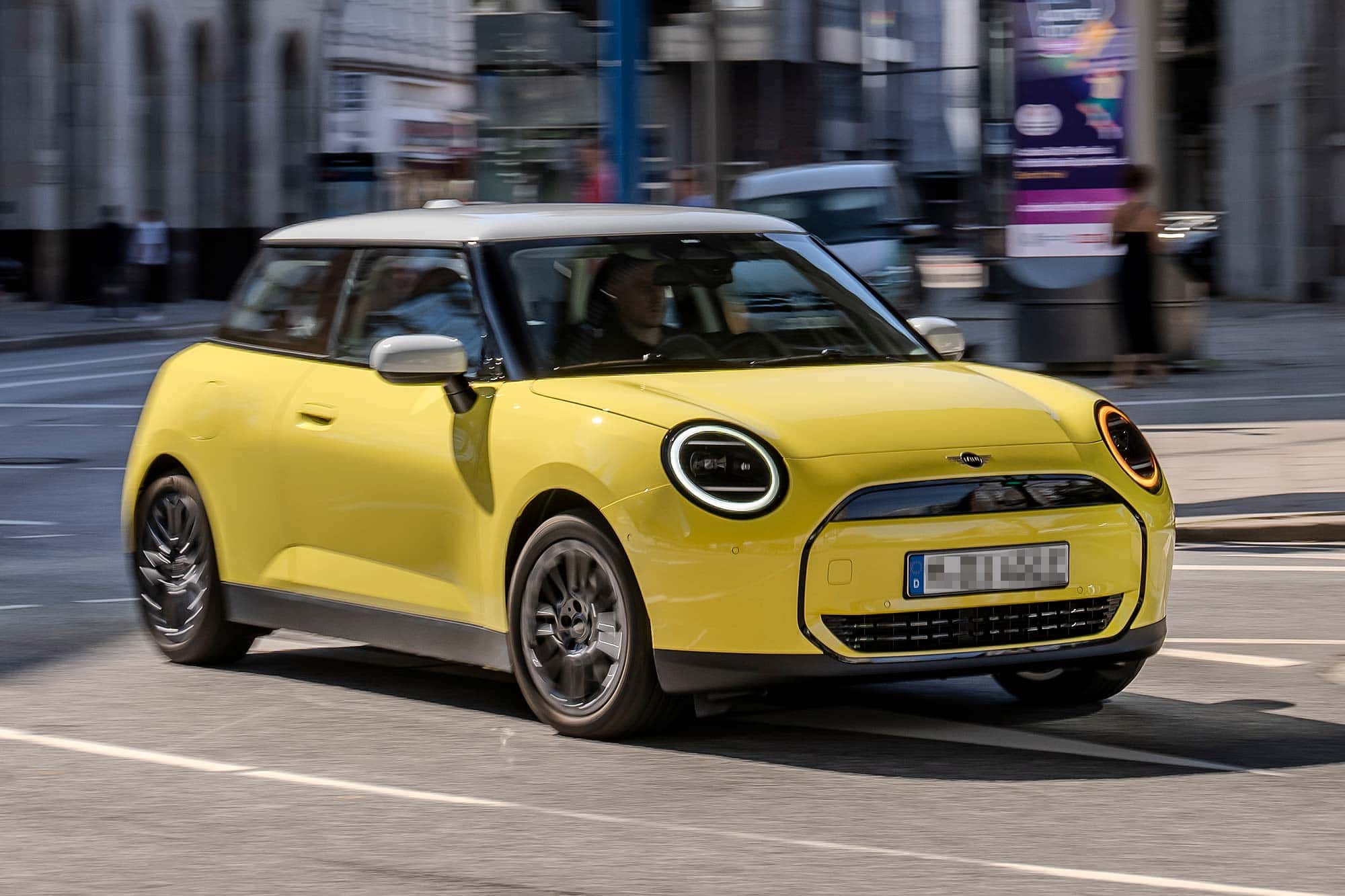 Yellow Mini Cooper with white roof driving on city street with blurred urban background