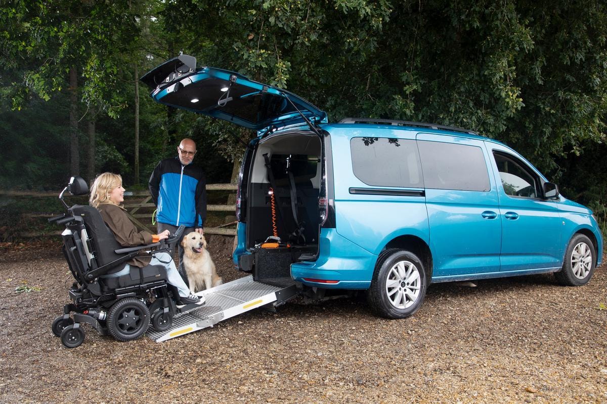 Woman in wheelchair with dog exiting blue accessible van via ramp, man standing nearby in wooded setting.