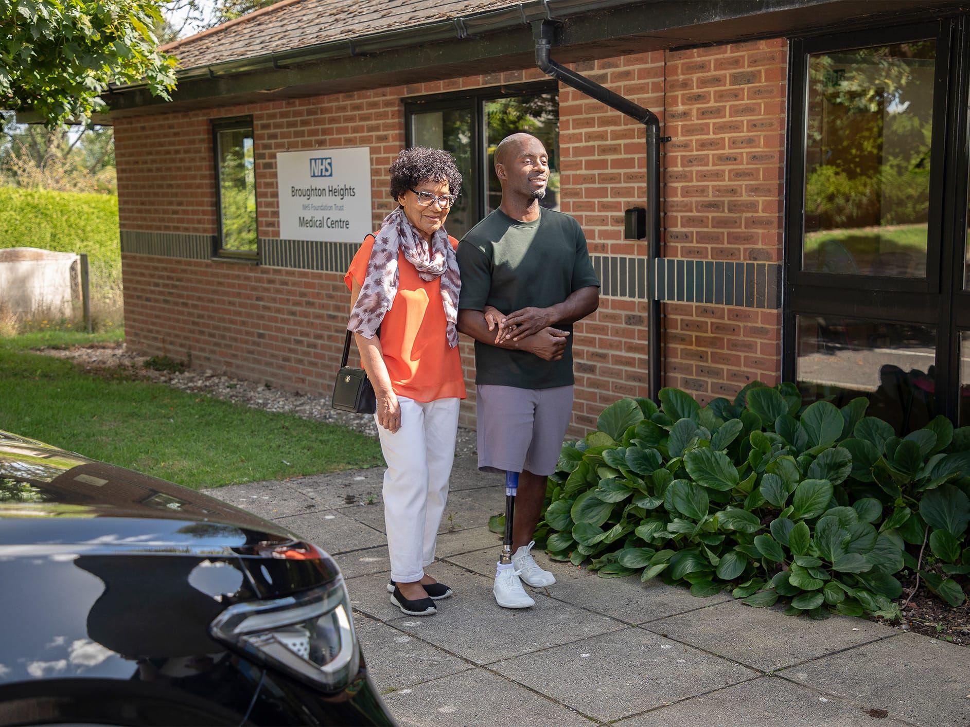 Two people leaving an NHS medical centre, with a woman in an orange top and man with prosthetic leg walking together.
