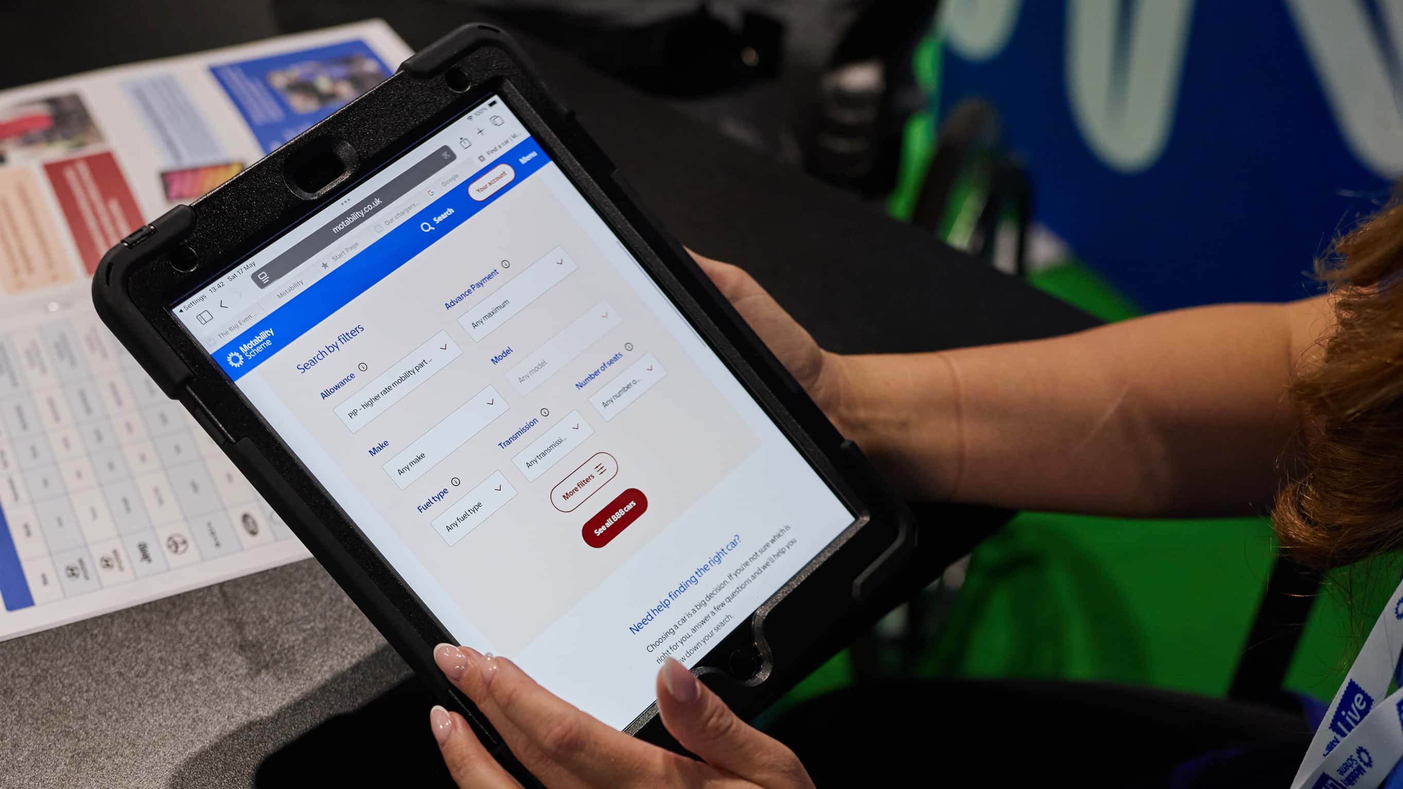 A person uses a stylus to navigate a Motability Operations webpage on an iPad. The screen shows a section titled “Check if you can get a car” with a button labelled “Start now”. The hand is tapping the screen, and the background is softly blurred.