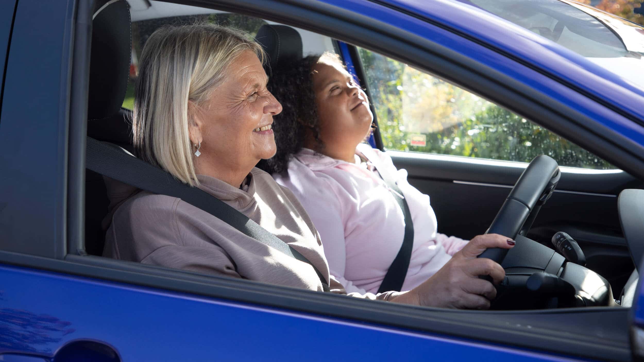 Two women sit in the front of a blue car on a sunny day. The driver smiles while holding the steering wheel and the passenger looks upward with a relaxed expression. Sunlight shines through the windows and greenery is visible outside.