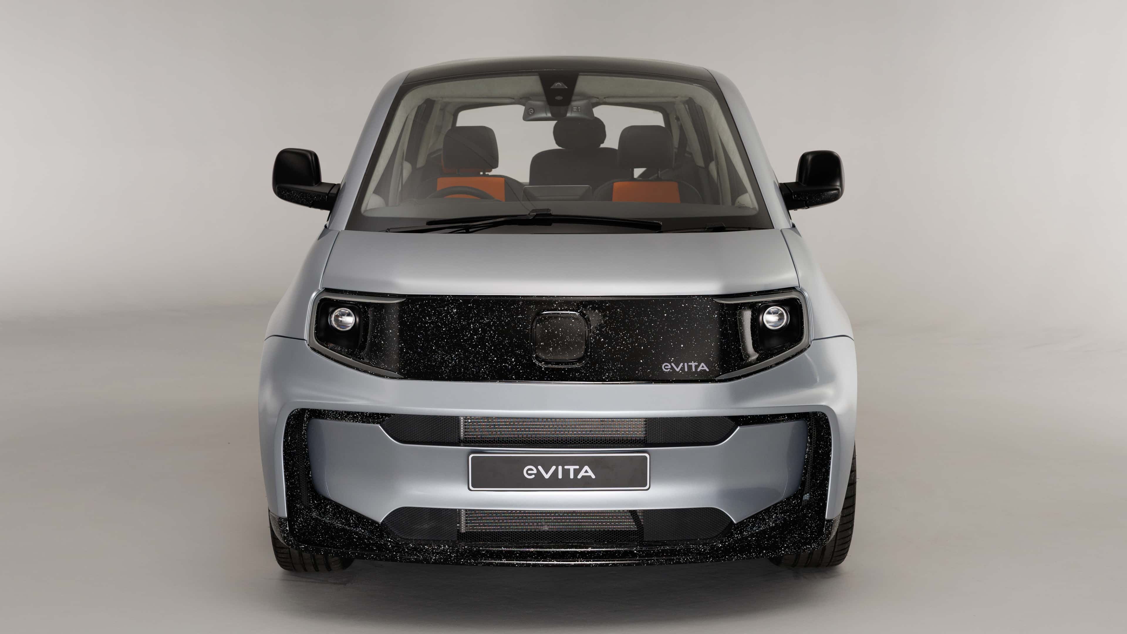 Front view of the e-VITA. The car features a smooth silver body with a black speckled front panel and integrated headlights, photographed in a studio setting.