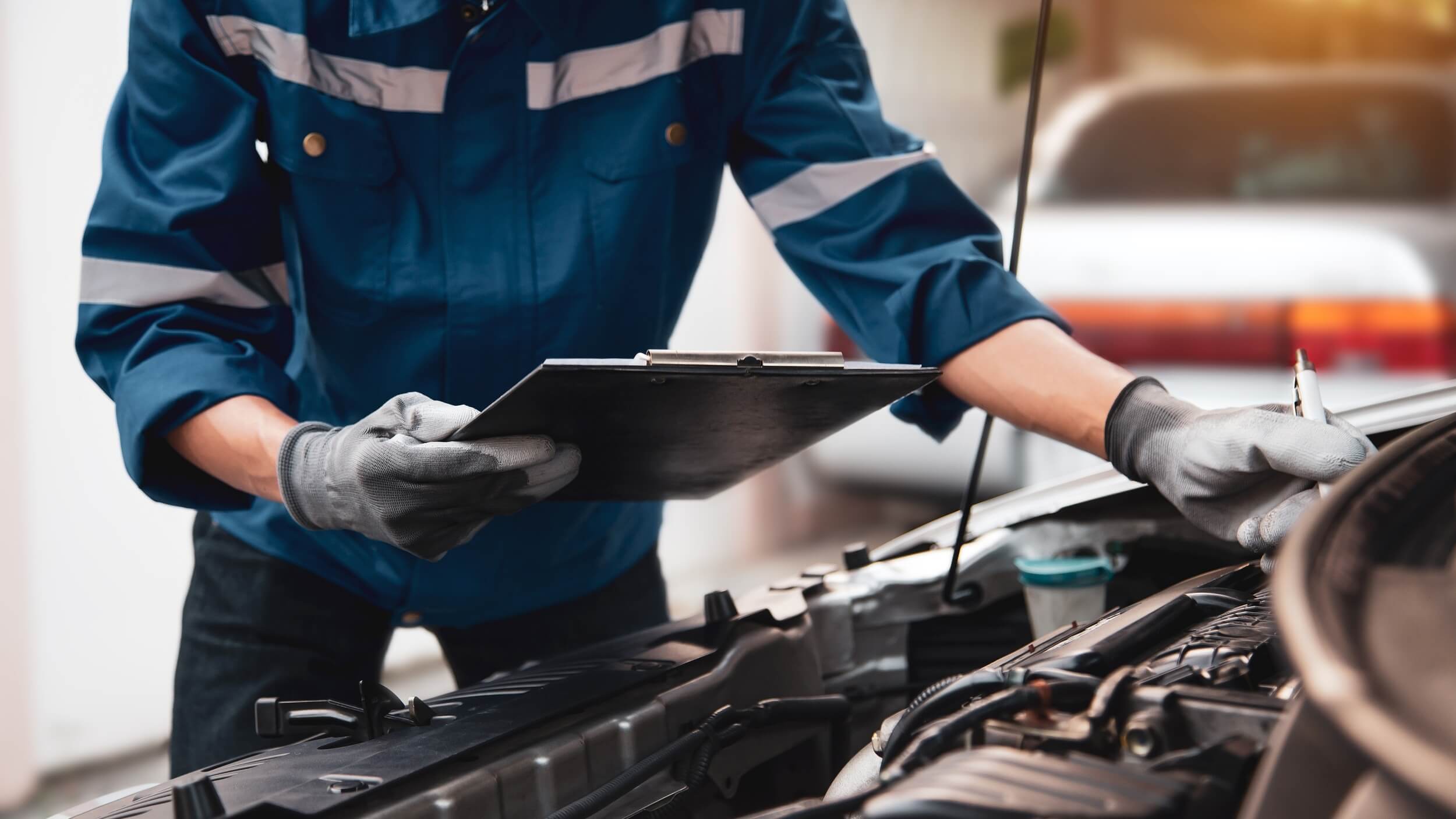 Mechanic in a blue work uniform and gloves inspecting a car engine while holding a clipboard, with the bonnet open in a garage setting.