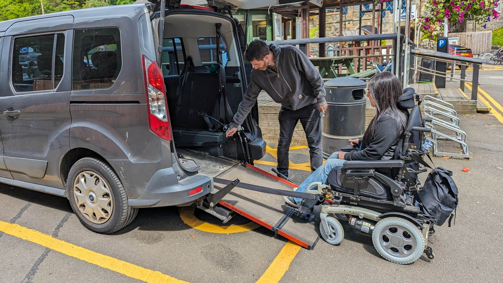 A man lowers a ramp from the rear of a grey Wheelchair Accessible Vehicle (WAV) while a woman using a powered wheelchair waits at the bottom of the ramp in a car park with buildings and bike racks in the background.