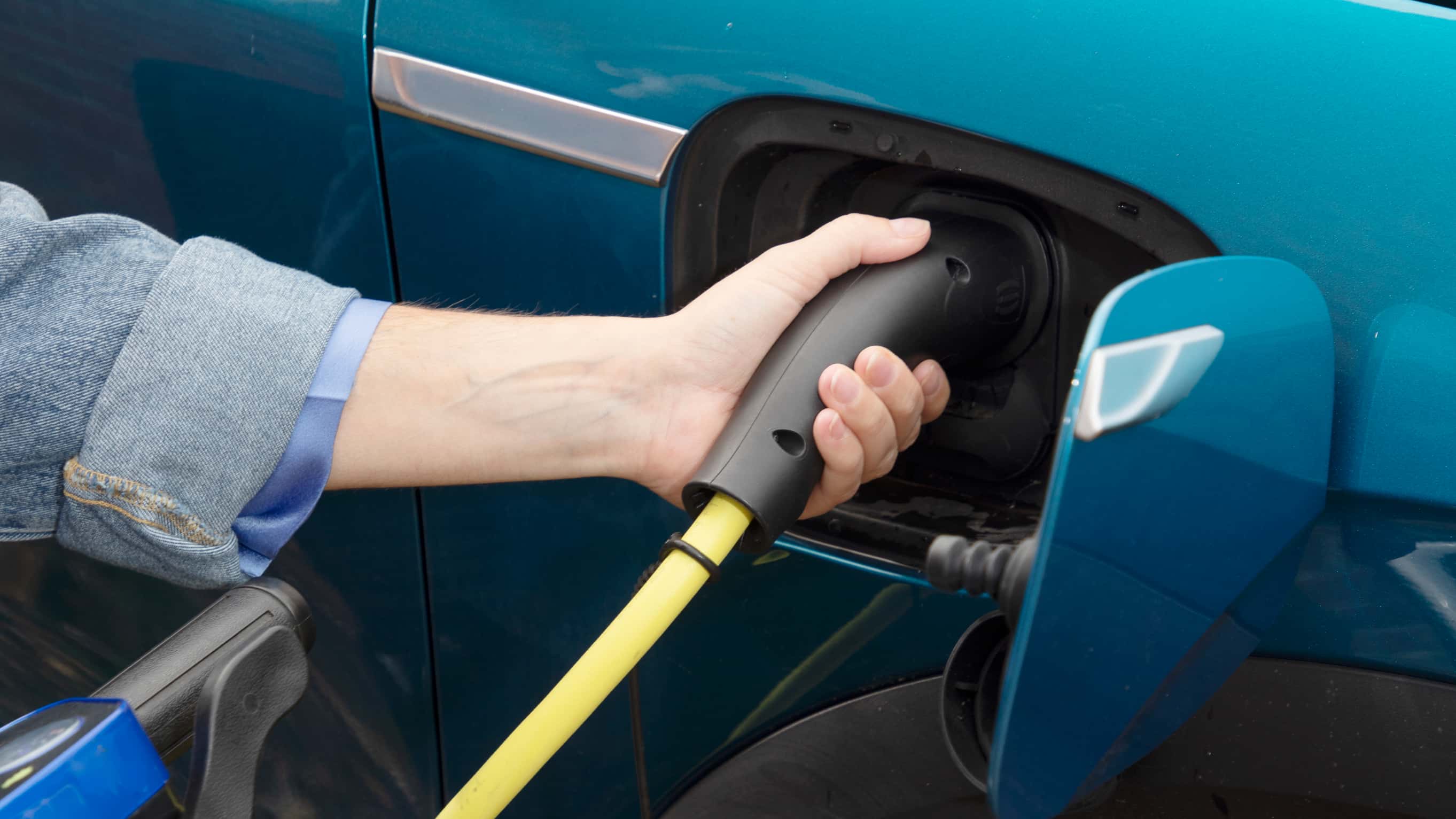 Close-up of a person’s hand plugging a yellow electric vehicle charging cable into a blue car.