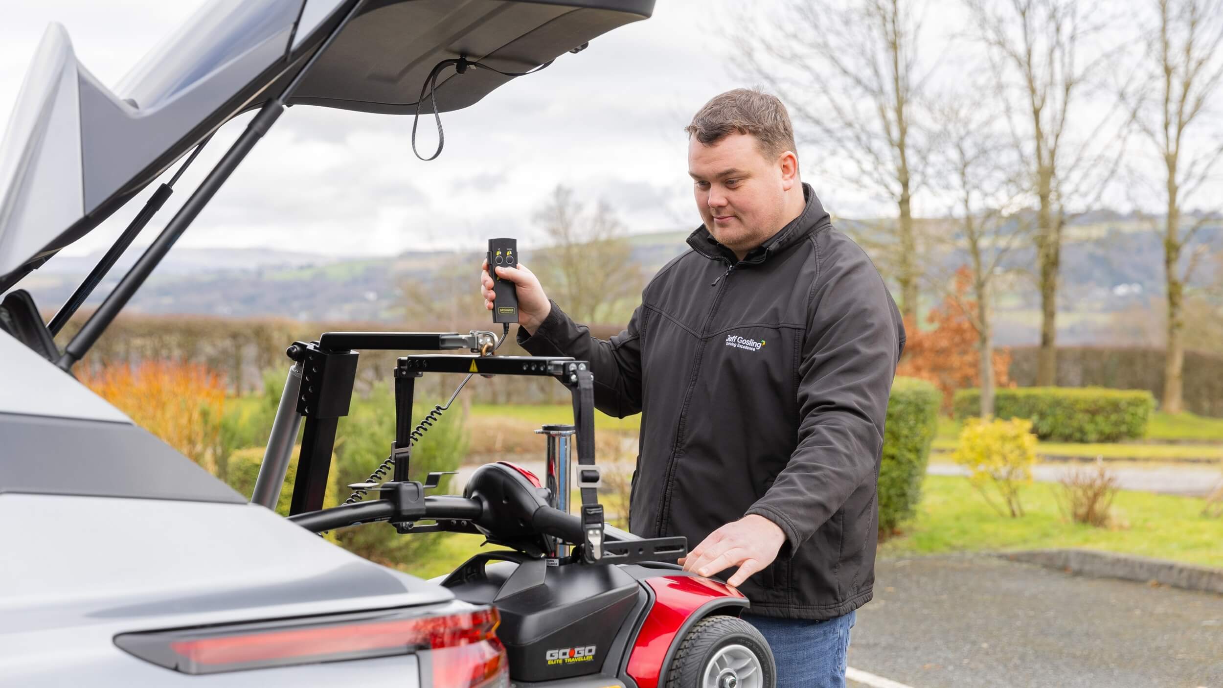 A man operating a Jeff Gosling electric hoist to lift a red mobility scooter into the boot of a grey SUV. He stands beside the open boot, holding the hoist’s remote control, with trees and countryside in the background.