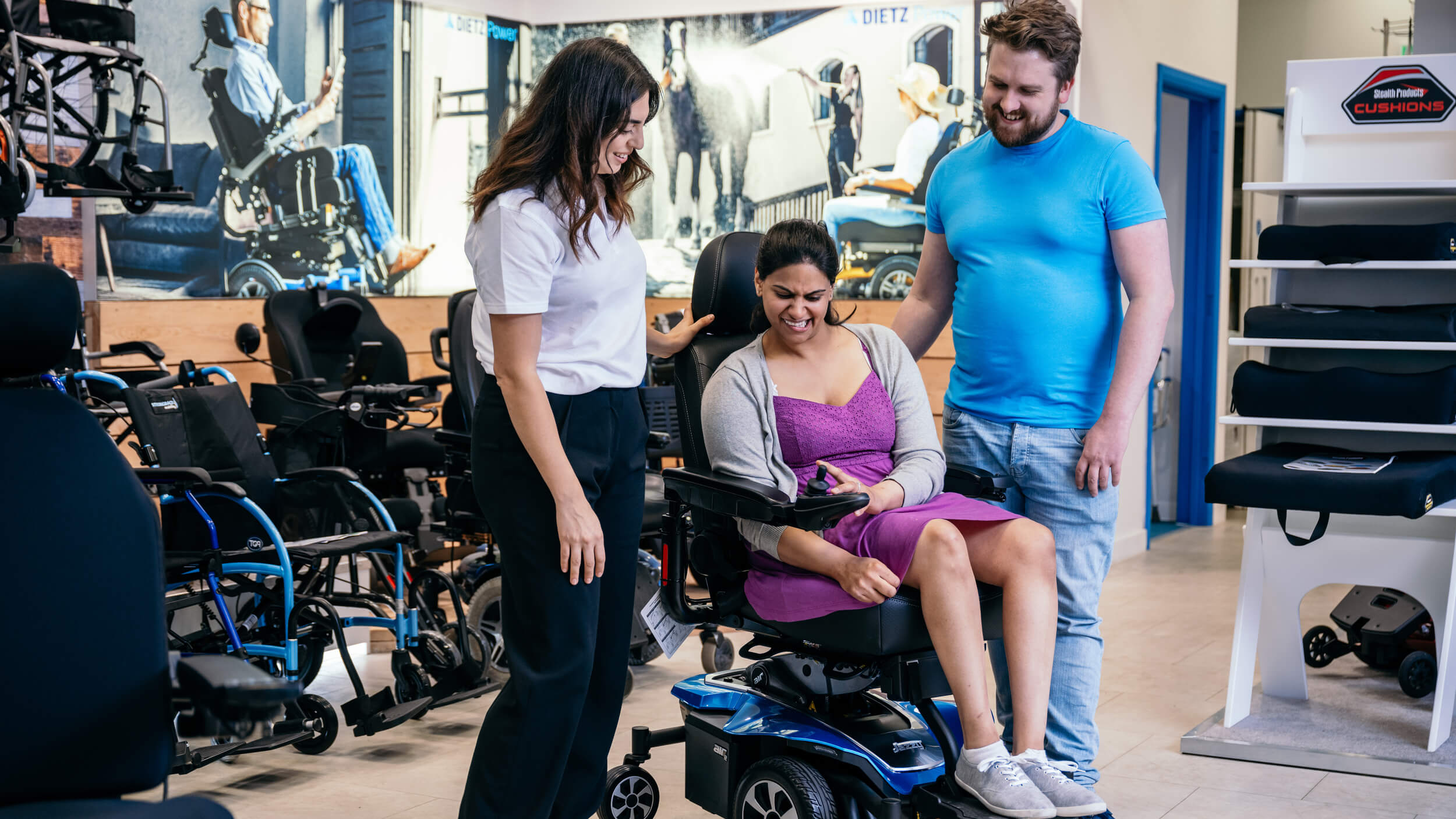 A smiling woman tests a powered wheelchair in a mobility showroom, assisted by a staff member and a man standing beside her. Various mobility aids are displayed around them.