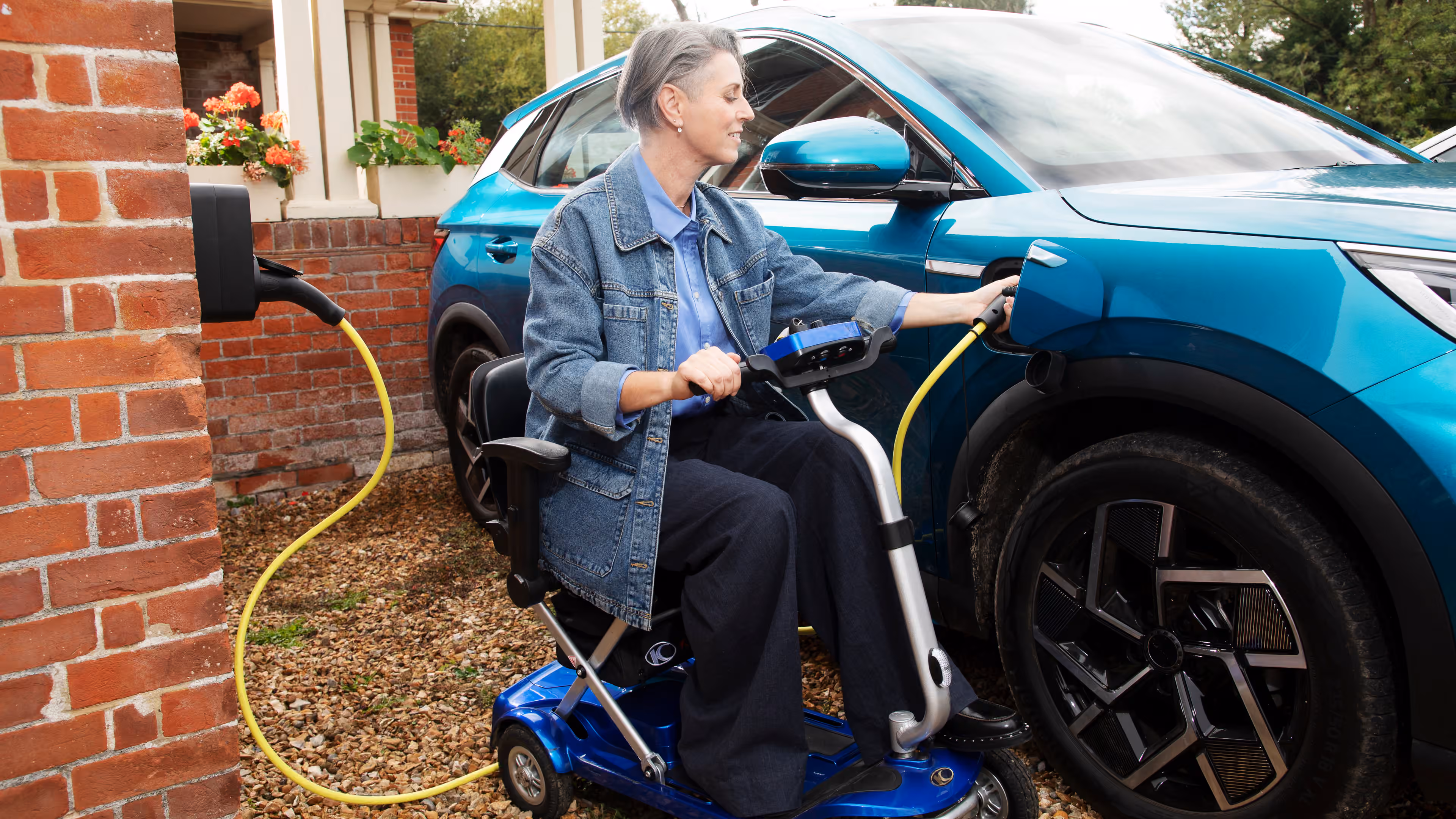 A person using a mobility scooter plugs a charging cable into a blue electric car outside a home.