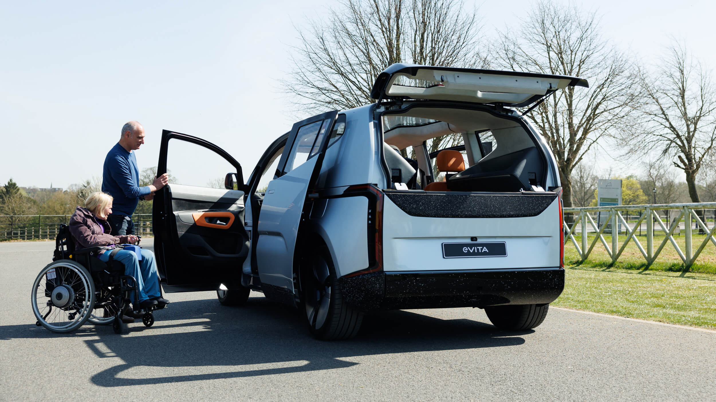 A man assists a woman in a wheelchair beside an eVITA electric vehicle parked outdoors. The car’s rear and side doors are open, showcasing its accessible design with a spacious interior and vibrant orange accents. The setting is a bright day in a park-like environment.