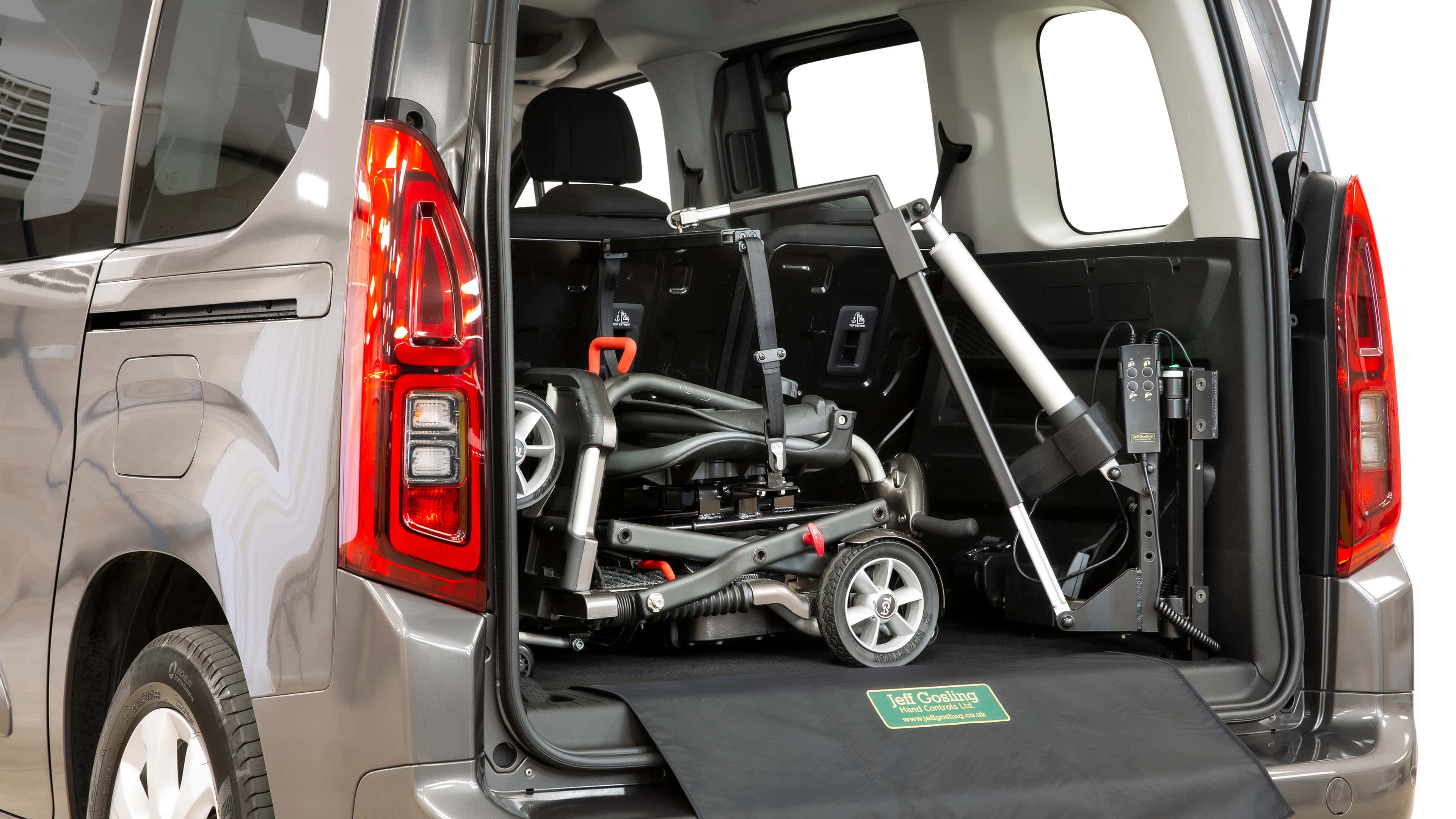 A folded mobility scooter is secured in the back of a grey vehicle using an electric hoist. The hoist arm extends from the boot floor, with straps and controls visible, and the boot ramp displays a Jeff Gosling Vehicle Adaptations logo.