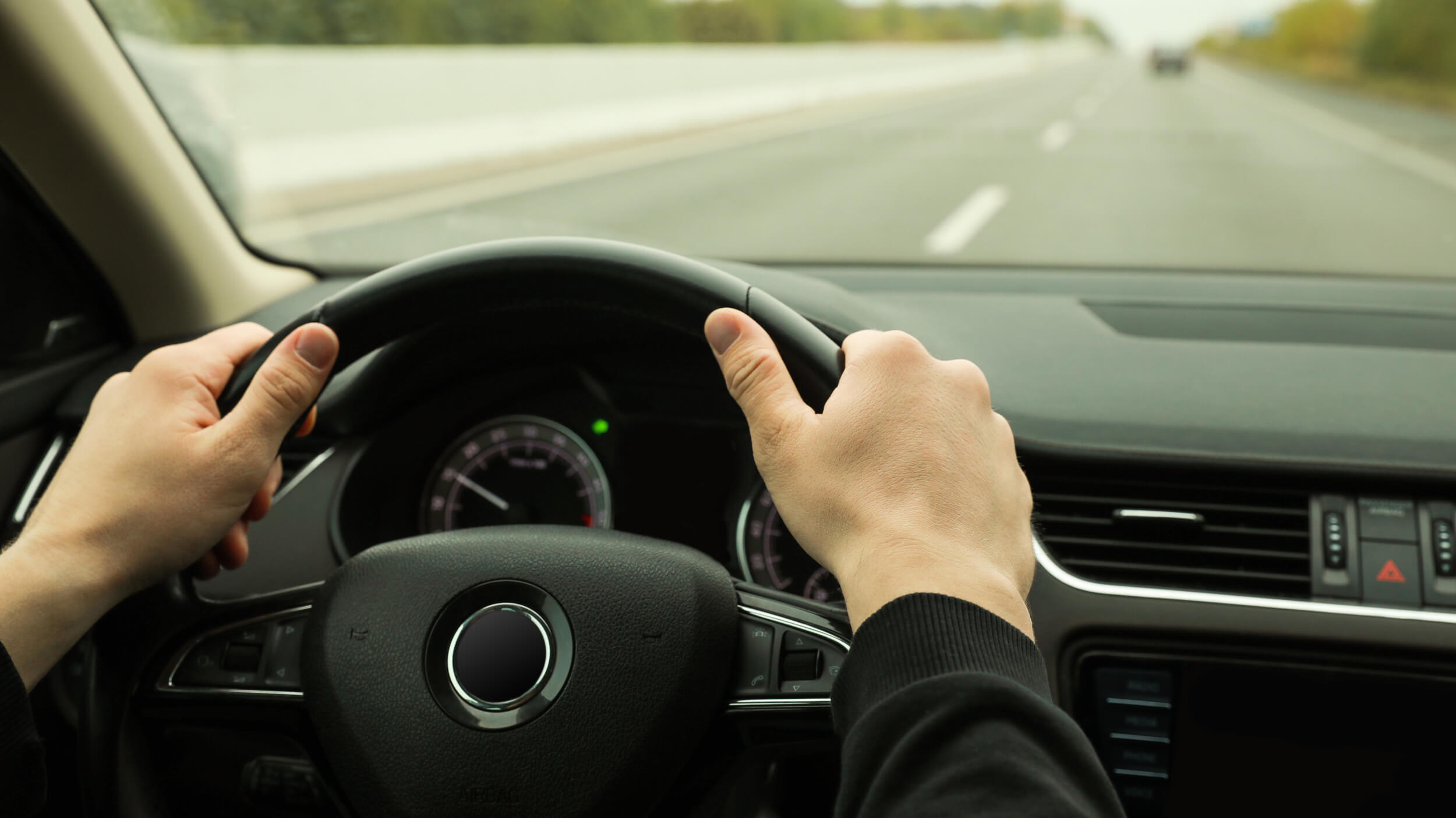 Driver holding a steering wheel on a quiet road, viewed from inside the car.