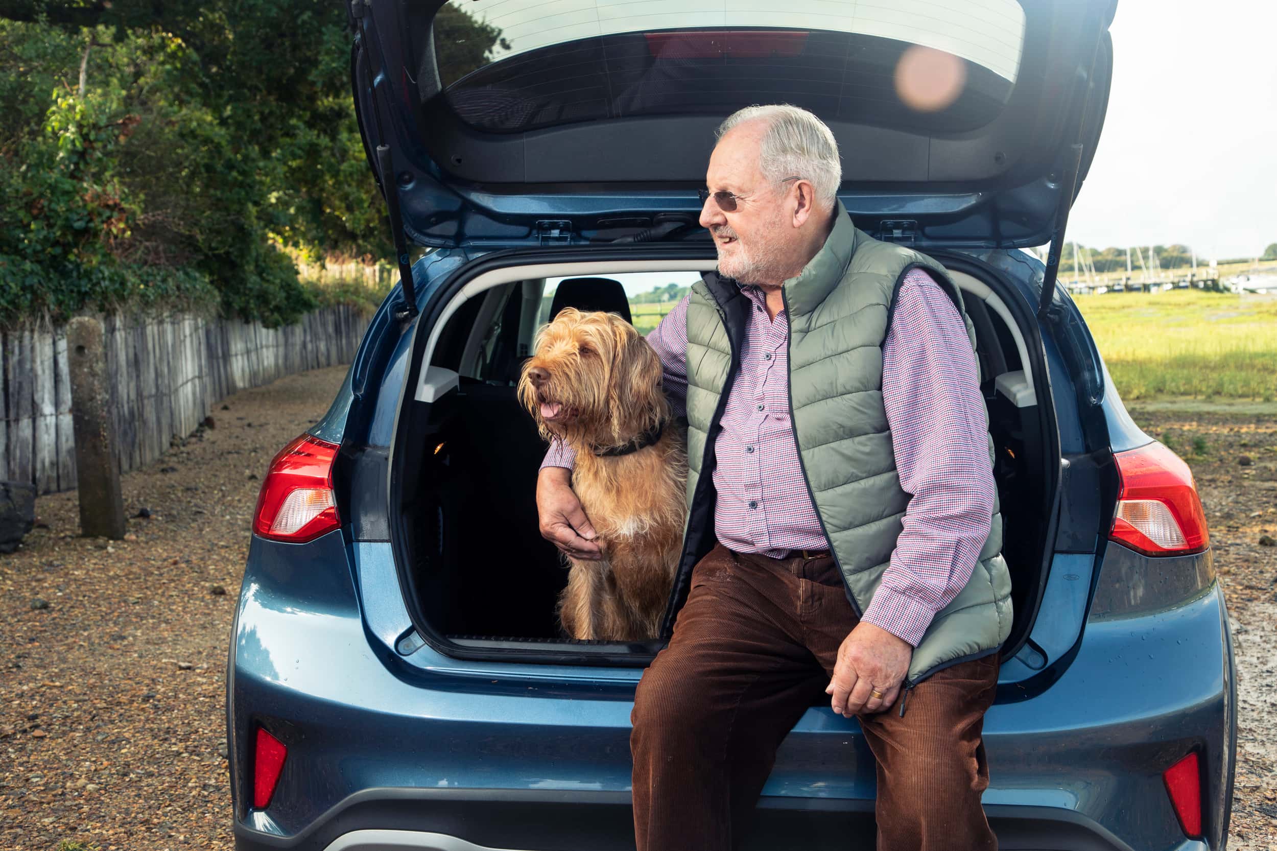 An older man sits on the boot of a blue car with his dog beside him.