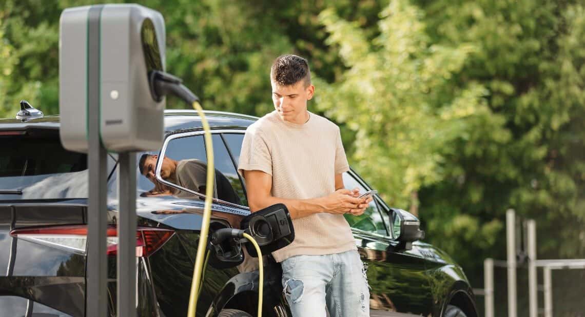 Men charging electric car in city and adjusting EV charging app on smartphone in hot weather
