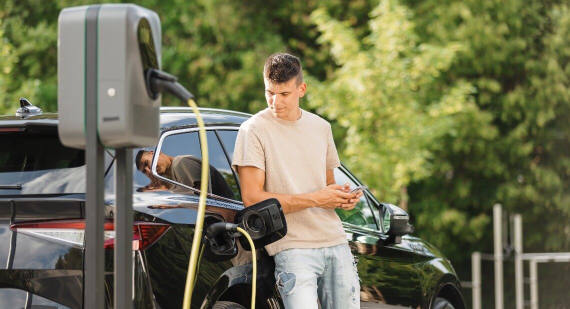 Men charging electric car in city and adjusting EV charging app on smartphone in hot weather