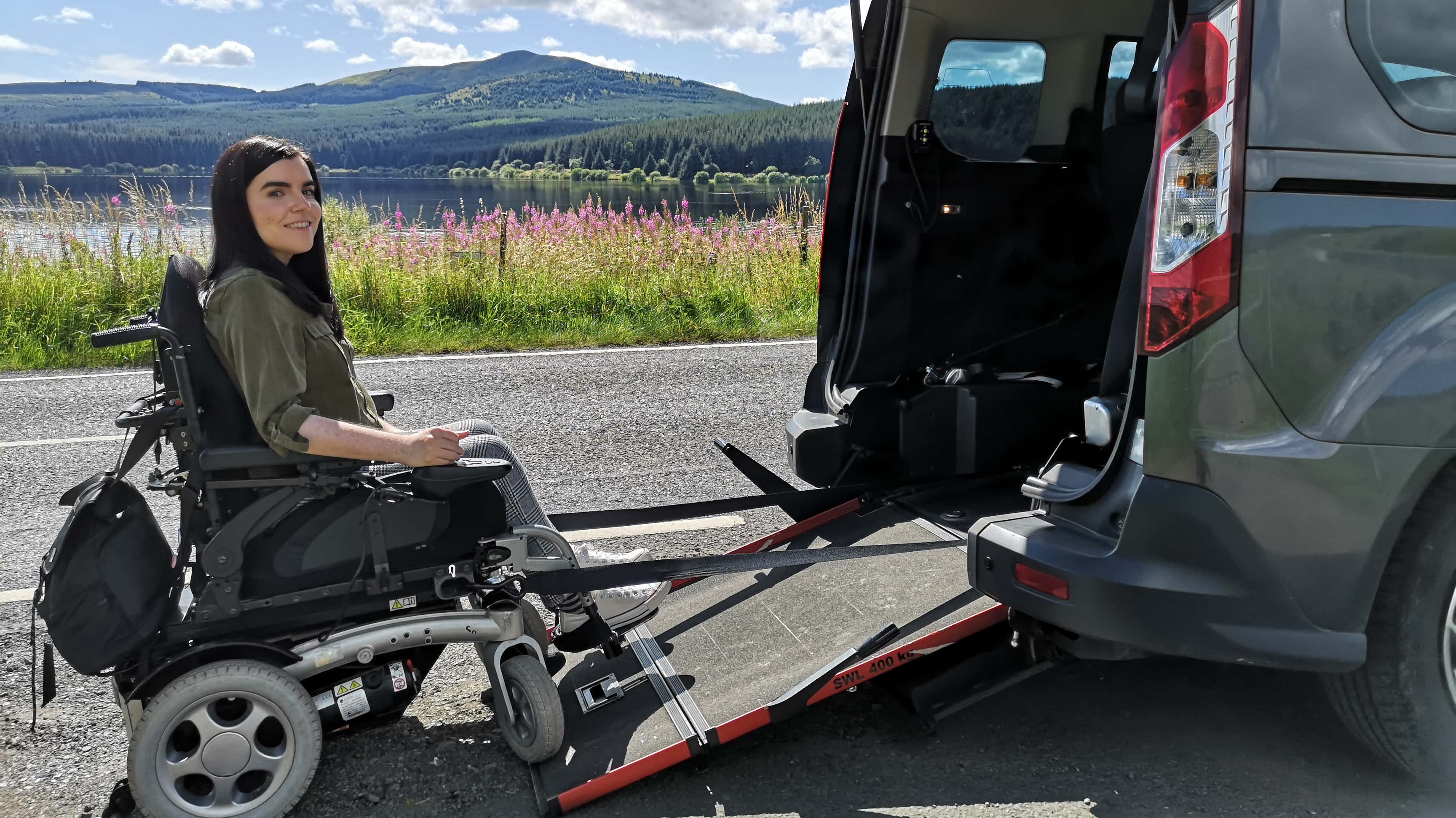 Emma Muldoon sits in a powered wheelchair beside a Wheelchair Accessible Vehicle (WAV) with a rear ramp deployed, parked by a lake with hills and trees in the background on a sunny day.