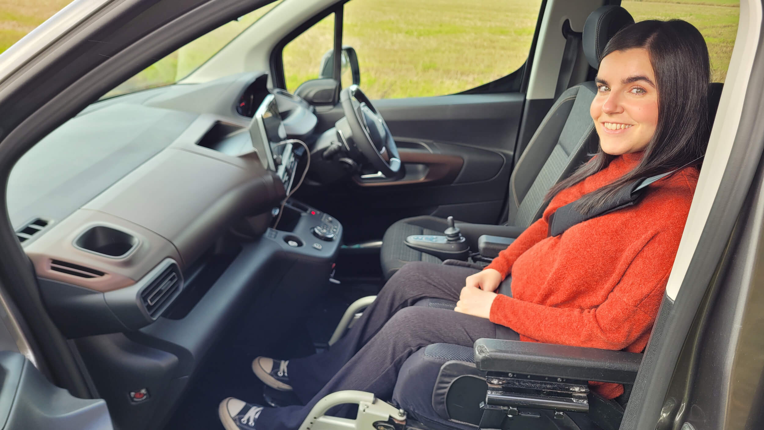 Emma Muldoon smiles while seated in the front passenger position of a WAV, secured in her powered wheelchair inside the adapted vehicle.