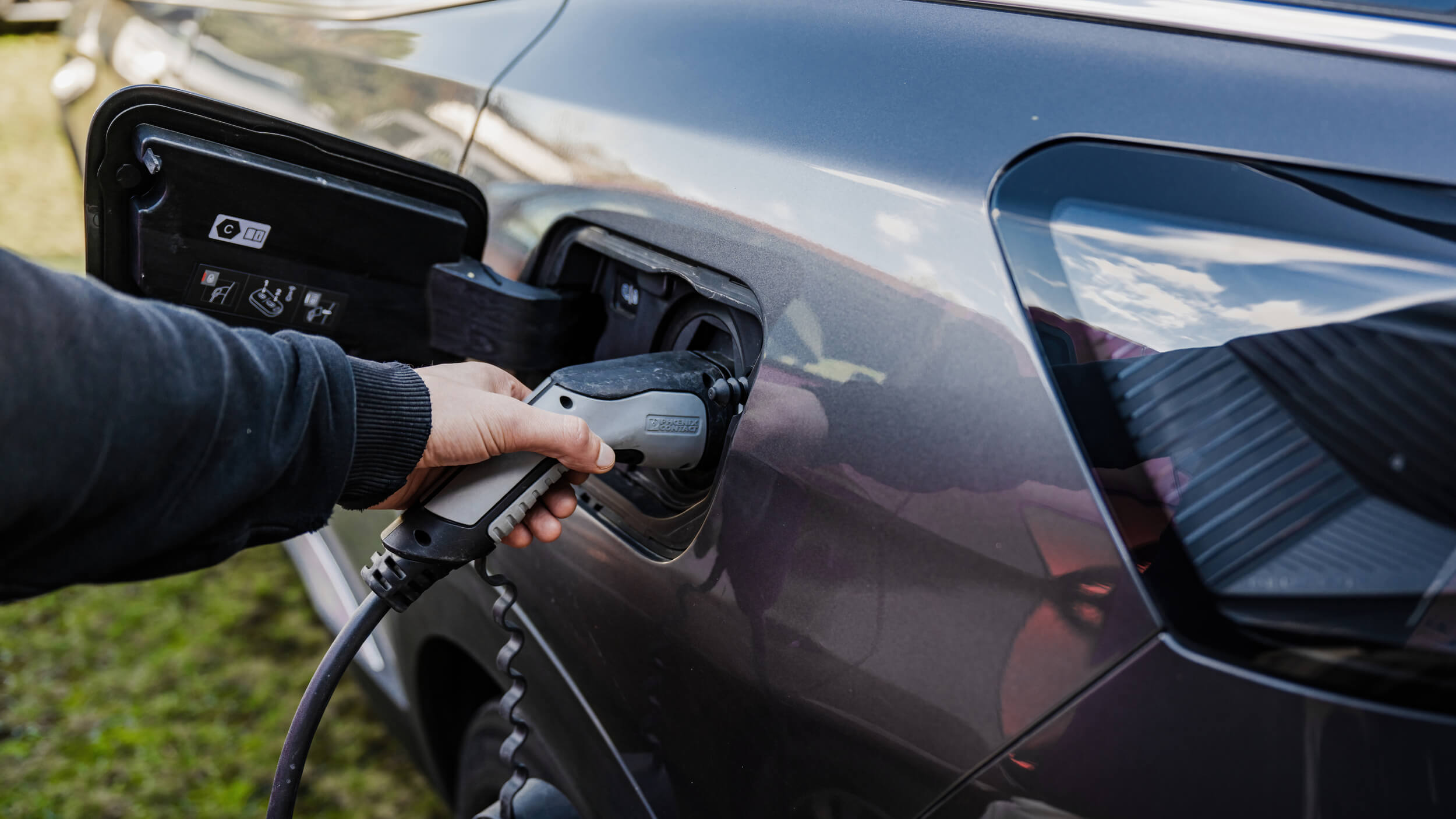 A close-up of a person’s hand plugging an electric vehicle (EV) charging cable into the charging port of a dark grey car, with grass and sky reflected in the car's surface.