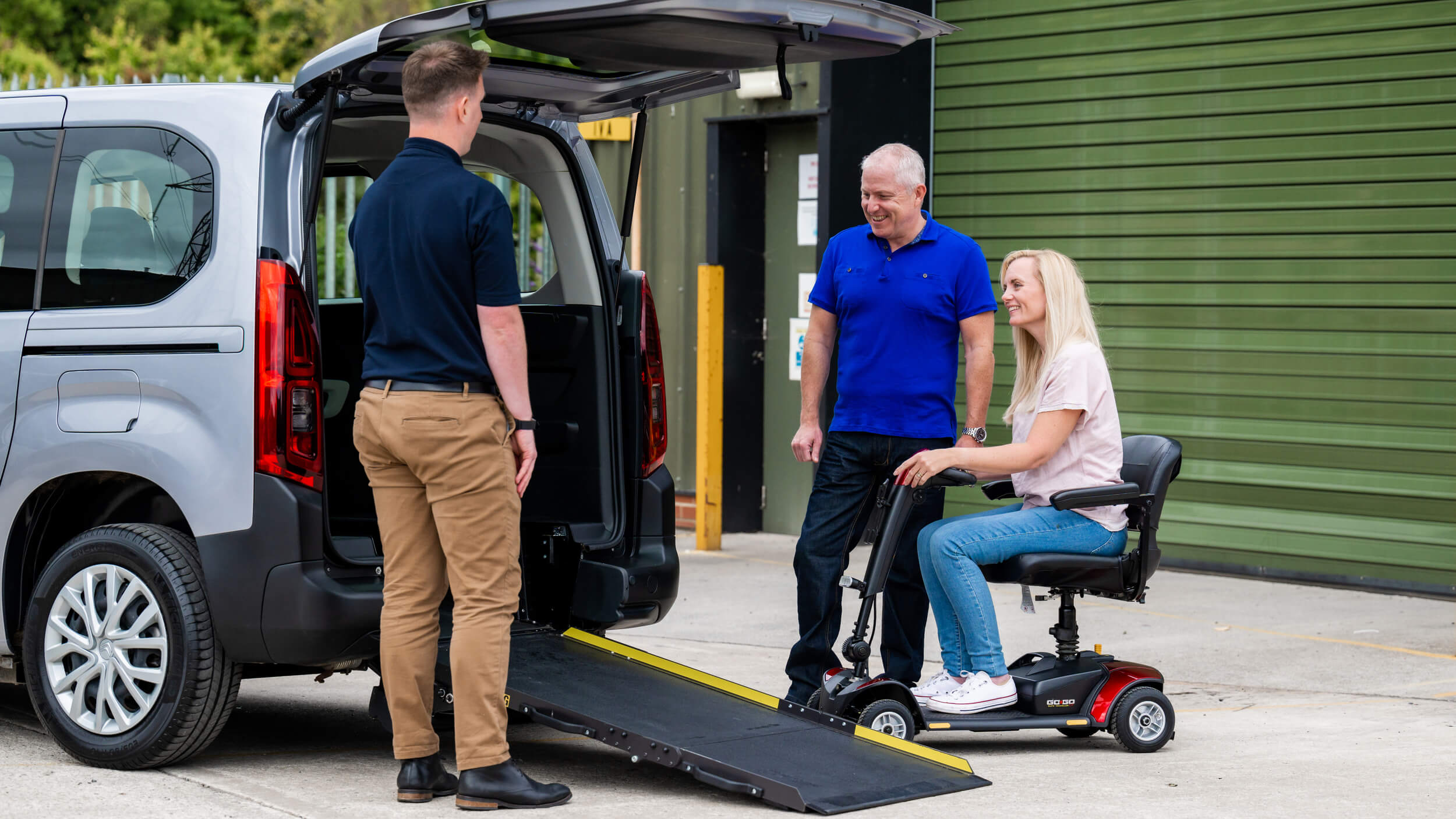 A woman on a red mobility scooter smiles as she approaches the rear ramp of a wheelchair accessible vehicle (WAV), while a man in a blue shirt and a mobility advisor stand nearby offering support.