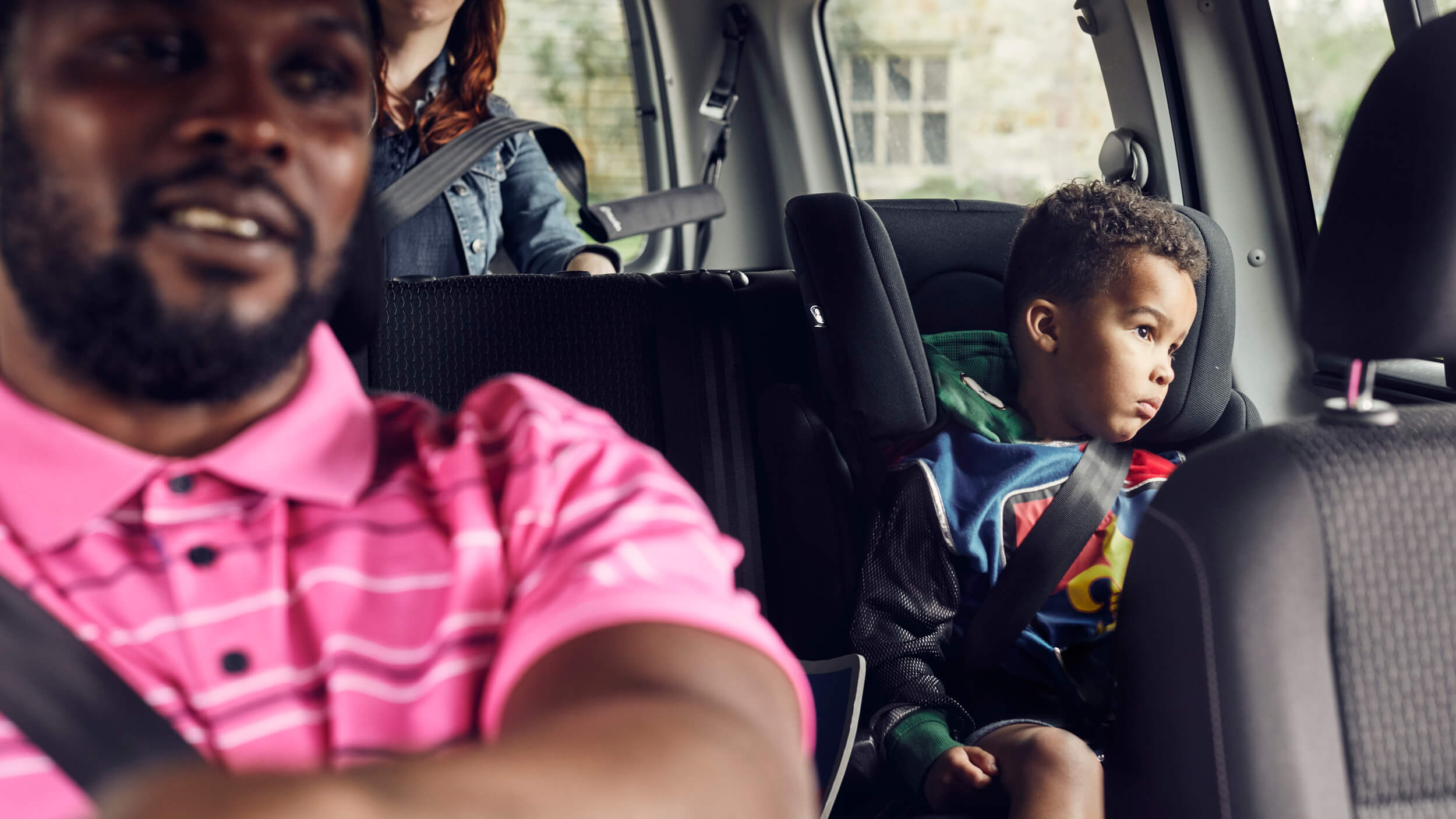 A man drives a car with a woman in the back seat and a young child sitting safely in a car seat, secured with a seatbelt and looking out the window.