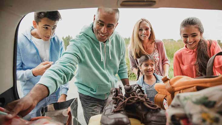 A family of five happily unpacks or packs the boot of their car for a trip. The father, wearing a green hoodie, reaches in to arrange bags and hiking boots, while the mother and three children smile and look on. The background shows an outdoor setting wit
